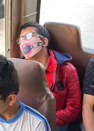 一名男子在公共交通工具（可能是公交车）上睡着了，他穿着红色外套和蓝色连帽衫，戴着眼镜。最引人注目的是他嘴上戴着一个搞怪口罩，这个口罩设计成夸张的张开嘴巴造型，与他沉睡的状态形成滑稽对比。周围还有其他乘客坐在相邻座位上。