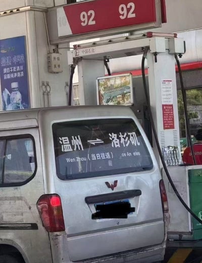 这张实拍图拍摄于中国石化加油站的92号汽油加油机旁，一辆银灰色五菱宏光面包车正在加油，车辆后窗上贴有一张趣味恶搞贴纸，贴纸用双向箭头连接“温州”与“洛杉矶”两地，还标注了“当日往返”的字样，搭配拼写错误的英文文本，用极度夸张的方式玩“五菱宏光是神车”的网络梗，调侃该车型性能强悍、用途广泛，甚至可以实现跨大洲当日往返，笑点十足。