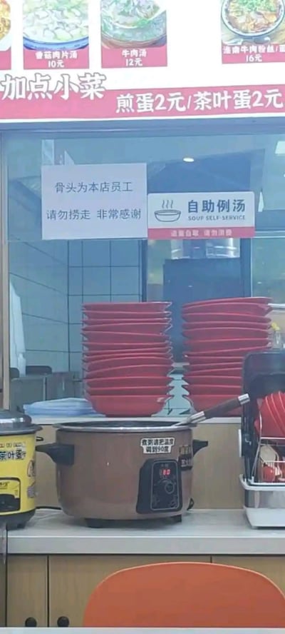 这是一张餐馆柜台区域的实拍照片，上方菜单板标注了多种汤品价格：香菇肉片汤10元、牛肉汤12元、淮南牛肉粉丝汤15元，还有“加点小菜”区域显示煎蛋和茶叶蛋均为2元。玻璃隔断上贴有手写告示：“骨头为本店员工 请勿捞走 非常感谢”，旁边是“自助例汤 SOUP SELF SERVICE”的标牌。下方操作台上堆叠着多摞红色盘子，摆放着两个电饭煲（一个黄色，一个棕色带数字显示屏），台面上还有餐具架和一把橙色椅子。整体场景充满生活气息，其中“骨头归员工”的告示因内容直白意外显得有些幽默。
