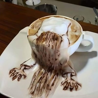图片展示了一杯放在白色方形盘子上的咖啡，杯中的奶泡被精心塑造成“倾泻”的视觉效果——白色奶泡混合深棕色咖啡液，从杯口向下流淌，在盘子上形成类似瀑布的纹理，仿佛咖啡正在“流泪”。背景为木质桌面，隐约可见金属容器和玻璃杯等模糊物体，整体构图聚焦于咖啡杯的创意拉花，视觉冲击力强。这是一种利用奶泡和咖啡液分层创造的错觉艺术，无文字或人物元素。