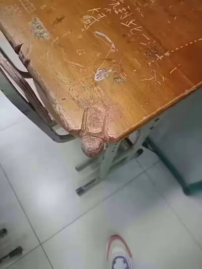 这是一张拍摄于教室中的破旧木质课桌的照片。课桌的角落被磨损和破坏，意外地形成了一个极具暗示性的造型，很像男性生殖器，因此带有强烈的恶搞和双关效果。桌面上刻有学生涂鸦和笔刻文字，充满了校园生活的气息和岁月痕迹。

桌角的特殊形状，常被网友用来整活或做表情包，属于“越看越不对劲”类型的校园恶作剧梗图。