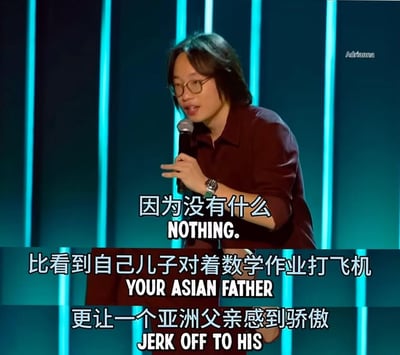 图片显示一位戴眼镜的长发男性喜剧演员正在舞台上表演脱口秀。他手持麦克风，背景是蓝色的舞台灯光。图片上有中文字幕和英文翻译，内容是一个关于亚洲父亲和儿子数学作业的笑话："因为没有什么比看到自己儿子对着数学作业打飞机更让一个亚洲父亲感到骄傲"。