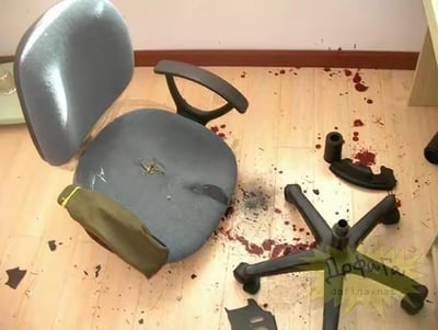 这张图呈现了一个看似惊悚的“意外现场”：一把灰色网布办公椅的椅身与底座支架分离，底座散落在浅色木地板上，地面和椅身沾染了大片仿血迹的红色痕迹，椅面有破损痕迹，还散落着黑色的塑料碎片，椅边放着一个军绿色小包。实际上这是一个恶搞恶作剧场景，利用仿血迹道具和破损的办公椅，模拟办公椅气杆爆炸“伤人”的意外，以此制造荒诞搞笑的效果，并无真实人员受伤。