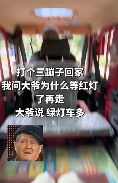 这是一张搞笑梗图，主体为从三轮机动车（三蹦子）后座拍摄的驾驶员背影，画面中央叠加了白色文字讲述段子：打三蹦子回家时，乘客询问开车的大爷为什么要等红灯的时候才走，大爷给出的回答是绿灯的时候车多。左下角插入了赵本山一脸疑惑、错愕的表情包，用来表达听到大爷这番反常理回答时的无语与不解，笑点在于大爷不符合常规认知的清奇出行逻辑，出乎意料又仿佛有一定道理。