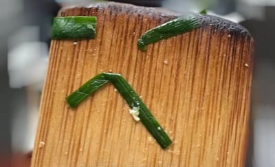 这是一张原始实拍的静态图，主体是一块带有使用痕迹的木质锅铲，其表面被人用三段绿色韭菜叶摆放出了拟人化的难过表情：两段韭菜分别作为向下耷拉的眉毛，一段弯折的韭菜形成向下撇的嘴角，让普通的厨房厨具拥有了低落的神态，充满生活趣味与创意感。图片底部带有一个包含数字的分享群水印。