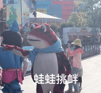 户外游乐园场景中，一只穿着蓝色围巾的红色斑点蛙蛙吉祥物正伸手指向旁边的蓝衣粉裙吉祥物，表情得意仿佛在挑衅对方。背景有色彩鲜艳的卡通风格建筑和树木，一名戴着黄色帽子和口罩的游客正在用手机拍摄这有趣的互动瞬间。图片底部配有文字'蛙蛙挑衅'，精准概括了画面核心。