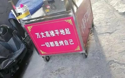 这是一张原始拍摄的静态标语图，画面主体是一个带滚轮的街头小吃推车，推车侧面设置了一块红色的装饰标语牌，牌上用白色字体写着“万丈高楼平地起 一切都是烤自己”，巧妙运用“靠”与“烤”的谐音，将原本励志的语句结合小吃摊的烧烤类经营属性进行改编，既贴合摊主的营生内容，又用诙谐调侃的方式传递出一种接地气的励志感。画面中还能看到推车的其他设备部分、旁边的电动车以及水泥质地的地面，整体场景是典型的街边摆摊环境。