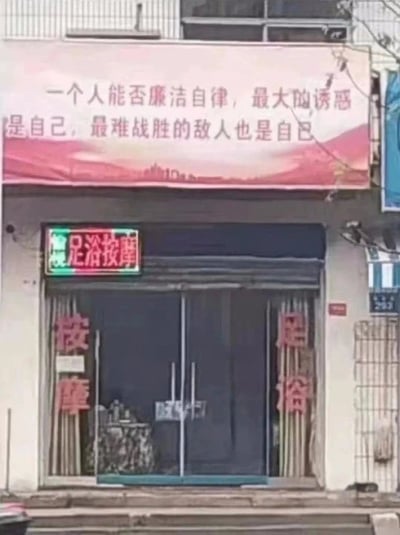 一张街头实拍照片，展示了一家足浴按摩店的正面。店铺上方悬挂着一条红色横幅，上面写着宣传廉洁自律的标语："一个人能否廉洁自律，最大的诱惑是自己，最难战胜的敌人也是自己"。店铺门口有霓虹灯招牌显示"足浴按摩"，两侧还有红色对联写着"按摩"和"足浴"。标语内容与店铺性质形成了有趣的对比，具有一定的讽刺和幽默效果。