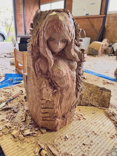 这是一张在工作室场景中拍摄的手工木雕作品实拍图，主体是一件尚未完成的女性形象木雕：木雕保留着部分原木树皮和粗犷的凿刻痕迹，面部被精细打磨出沉静柔和的神态，身体部分还带着明显的工具凿削纹理，呈现出一种被包裹的蜷缩姿态。木雕周围的榻榻米垫上散落着大量木屑和凿子等木雕工具，背景是带有木质结构的工作室空间，能看到窗外的车辆和室内的杂物，整体呈现出手工木雕创作过程中的真实场景，既有原木材质的粗犷质朴，又有面部刻画的细腻柔和。