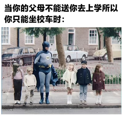 图片上方配有文字“当你的父母不能送你去上学所以你只能坐校车时：”，下方画面中，穿着经典蓝色蝙蝠侠制服的角色站在一群等待校车的儿童中间。背景是带有砖墙建筑和复古汽车的街道场景，蝙蝠侠表情严肃，与周围普通儿童形成鲜明反差，通过超级英雄身份与日常上学场景的对比制造幽默效果。