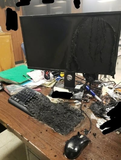 一张展示严重损坏电脑设备的照片。画面中央是一台电脑显示器，其右侧有大面积烧毁痕迹，塑料外壳明显熔化变形并呈黑色焦状。显示器前方的键盘同样遭受严重烧毁，整个表面被黑色烧焦物质覆盖，按键已无法辨认。桌面上散落着各种物品，包括绿色文件夹、纸张、电线和一个幸存的黑色鼠标。背景中可以看到办公环境的柜子和窗户，图片中有几处黑色涂抹的 censorship marks。