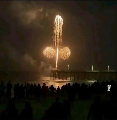 夜晚的海边，一群人聚集在岸边观赏盛大的烟花表演。夜空中绽放着三朵明亮的烟花，中间是一道高耸的金色光柱，两侧各有一朵圆形的金色烟花，形成独特的造型。下方是平静的海面和一座延伸至海中的码头，岸边挤满了观赏烟花的人群剪影，整体氛围充满节日的喜庆与热闹。