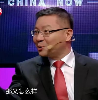 这是一张政论节目《这就是中国》的视频截图，图中人物为学者张维为，他身着深色西装、白色衬衫，搭配红色领带，佩戴着耳麦，侧头看向画面左侧，嘴巴微张，神态带着一丝从容与不在意。画面下方配有白色字幕‘那又怎么样’，背景上方有英文‘CHINA NOW’的字样。这张图常被制作成表情包，用于在网络聊天中表达对质疑、不同声音不在意，或是从容回应的情绪，没有双关、谐音梗等特殊含义。