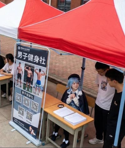 图片展示了大学社团招新现场，一个名为“男子健身社”的摊位格外引人注目。摊位上方是红白色相间的遮阳棚，旁边立着一块宣传展板，展板上印有多张肌肉发达的男性健身照片，下方配有多个二维码。摊位前的桌子后坐着一位蓝发女生，她穿着类似校服风格的制服，戴着贝雷帽，双手交叠放在身前，面带微笑。两名男生正站在桌旁，似乎在咨询社团相关信息。整个场景的反差感十足：以“男子健身”为主题的社团，却由一位造型可爱的蓝发女生负责招新，形成了有趣的视觉和主题对比。