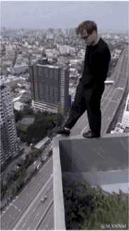 这是一段动态GIF，画面中一名男子身处高楼边缘，做出向另一处平台跳跃的动作，背景是车水马龙的城市道路与密集的高楼建筑群，看起来场面十分惊险刺激，但实际上这是利用视觉错位的拍摄手法营造出的假象，男子并没有处于真正的危险处境，只是拍摄角度让观众产生了错误的空间认知。
