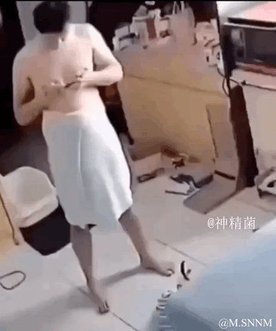 这是一段搞笑的动态视觉错位视频：画面里一名裹着白色毛巾的男子站在杂乱的室内，正低头摆弄胸前的物品。从初始视角看，毛巾下方仿佛有个狗头从他两腿间探出来，镜头拉近后才揭晓真相——这只是地面上的白色物品和拍摄视角共同造成的错觉，荒诞的反差感制造出十足的搞笑效果。