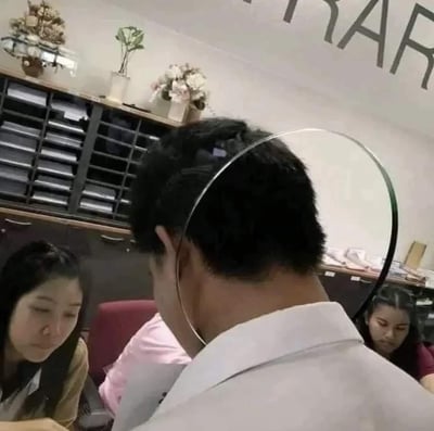 这张图拍摄于日常的办公室场景，画面中一位穿白衬衫的男士背对镜头，他身后的弧形金属条因为视觉错位，恰好和男士的头部位置重合，看起来像是从他头顶延伸出来的天使光环，让普通的办公室瞬间多了趣味感；画面里还有两位低头专注的女同事，整体场景日常又因为错位效果显得十分搞笑。