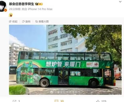 这是一张微博平台的发布内容截图，博主「暴食症患者李舜生」使用iPhone 14 Pro Max发布了一张实拍的厦门双层公交车照片。公交车身为绿色，印有啤酒瓶图案和宣传语「想炉管 来厦门」，这里运用了谐音梗，「想炉管」谐音「想撸串」，通过趣味谐音制造记忆点，用于本地宣传。背景是城市居民楼和绿树，天气晴朗，呈现出日常的城市街景。