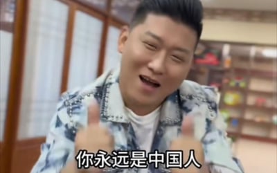 图片中一名男子穿着蓝白花纹夹克，身处带有木质框架和书架背景的室内环境中。他双眼微眯，嘴巴张开，面带笑容，双手竖起大拇指，表情显得开心且激动。图片底部有文字“你永远是中国人”，整体传达出强烈的身份认同和积极情感。