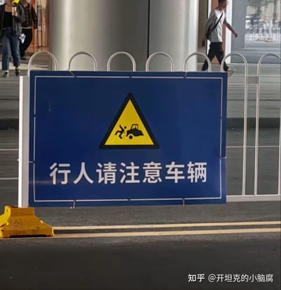 这是一张在城市街头拍摄的交通警示标志牌，标志牌主体为蓝色，上方配有黄黑三角形的车辆伤人警示图标，下方用白色字体标注文字“行人请注意车辆”，用于警示过往行人留意来往车辆，防范交通事故。背景中可见城市街道的护栏、路面以及路过的行人。