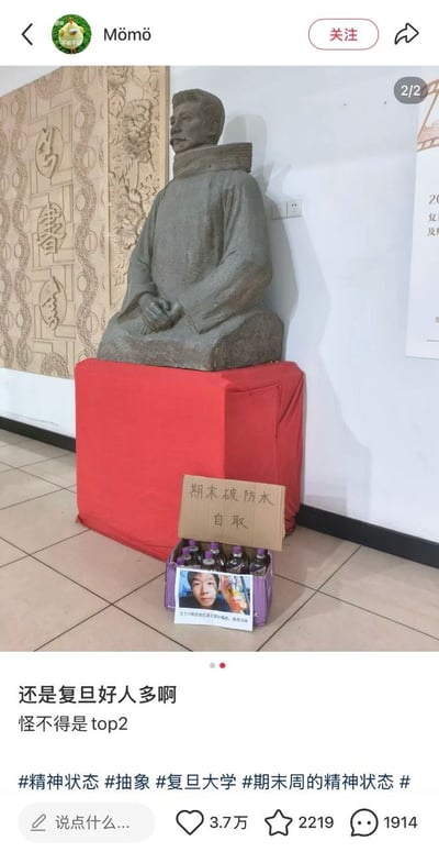 图片展示了复旦大学内一座历史人物雕像，雕像端坐于红色基座上，基座前放置着一个装有饮料的纸箱，上面贴着写有“期末破防水 自取”的纸板。这是学生们在期末周创造的幽默暖心之举，反映了校园生活中的轻松一面。