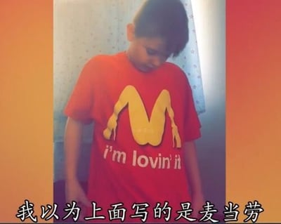 这是一张带文字注释的静态单图，主体是一名低头站立的真人男孩，他穿着一件红色恶搞T恤，T恤上的图案第一眼看起来是麦当劳经典的金色M金拱门标志，但实际上该图案被低俗改设计成了女性臀部加双腿的造型，图案下方还保留了麦当劳的品牌标语"i'm lovin' it"，图片底部配有中文配文"我以为上面写的是麦当劳"，调侃第一眼会将这个低俗设计错认成普通麦当劳logo，依靠反差制造恶搞笑点。