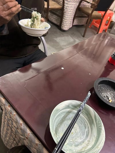 这张图片拍摄于一个朴素的用餐环境，展示了一位老人正在用筷子夹起面条进食，而前方桌上的碗已经空了，只剩下一点残渣和一双放好的筷子，有一种“吃饭比赛”的即视感。画面传达出了饭桌上的轻松幽默氛围，也让人联想到谁吃得快、谁先吃完的暗中较劲。