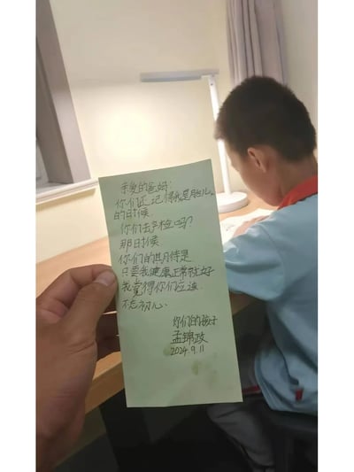 这张图片是家长视角的真实拍摄，画面中一只手拿着孩子孟锦政写的浅绿色便签，便签上孩子以自己还是胎儿时，爸妈产检时只希望自己健康正常的期待为切入点，委婉提醒爸妈“不忘初心”；背景里穿着浅蓝色校服的孟锦政正坐在书桌前专注写作业，整个场景温馨又充满细腻的情感，孩子用温柔的方式表达了对家长可能过高期待的回应，展现出孩童的敏感与懂事。