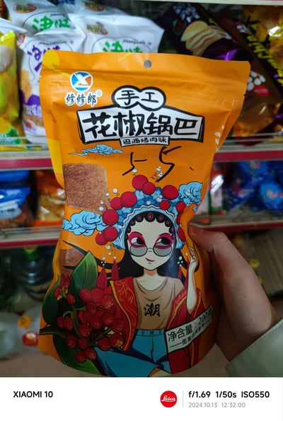 这是一张在超市货架前拍摄的修修郎手工花椒锅巴（巴西烤肉味）的商品包装实拍图，包装主色调为明亮的橙色，采用国潮风格设计，印有戴眼镜的戏曲风卡通女孩形象，女孩身着混搭潮服，搭配花椒图案与祥云元素，充满潮流个性。包装上清晰标注了产品名称、口味、净含量200克以及生产日期2024/01/05，背景货架上还能看到其他品牌的零食产品，整体场景展现了超市的零食售卖环境。