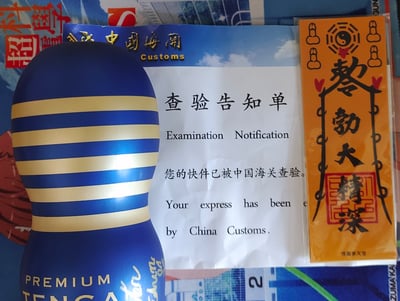 图片展示了一个蓝色和金色条纹的Tenga产品，旁边放着中国海关的查验告知单，以及一张写有'勤勉大精深'的黄色护身符。这种有趣的物品组合创造出一种幽默效果，暗示这个成人用品在海关查验时需要护身符的'保护'。