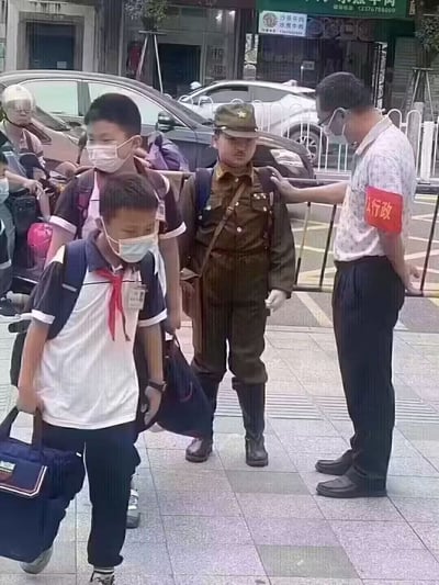 这是一张拍摄于小学门口的实拍照片，画面中有多名背着书包的小学生正准备入校，其余学生均穿着普通校服、佩戴口罩，其中一名男生没有穿校服，而是身着全套带有五角星帽徽的旧式棕色日本军装，搭配同色系长靴、白手套，还斜挎着帆布包，他被一旁佩戴印有“行政”字样红色袖章的学校工作人员拦住，工作人员的手搭在该男生的肩膀上，似乎正在对他进行劝阻或询问，背景可见临街的商铺招牌、停放的私家车与道路隔离护栏。