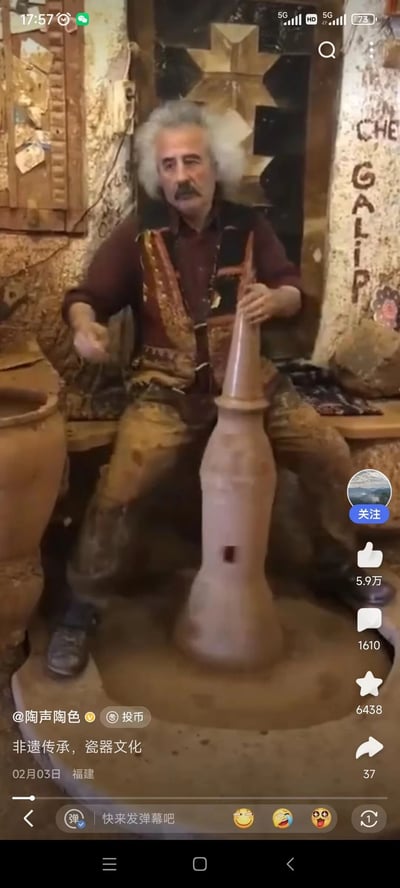这是一张哔哩哔哩平台的视频截图，画面中一位外形酷似爱因斯坦的老年男性陶艺匠人，身着带有民族特色的服饰，正坐在充满复古质感的陶艺作坊内，通过脚动方式驱动陶艺转轮，双手专注地对转轮上的陶坯进行塑形。作坊的墙面和陈设都带着手工制陶场所的质朴与厚重感，视频底部配文“非遗传承，瓷器文化”，展现了传统手工陶艺的制作过程，传递出非遗文化的独特魅力。