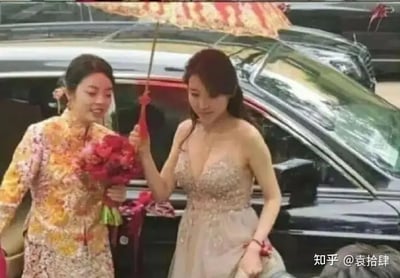 图中是一场婚礼迎亲的场景，一名身着精致中式刺绣旗袍，手捧花束的女性与另一位穿着低胸礼服的女性一起在车辆旁行走，并撑着一把中式油纸伞。画面展现了中西结合的婚礼气氛，有传统与现代元素的融合。图片氛围热闹，可能是在婚礼仪式中的某个环节，如接亲或新娘出门时刻。整体风格洋溢着喜庆与热烈的情感，非常适合形容“中西结合”、“氛围感拉满”或是婚礼文化差异的社交网络话题。

未见明显双关或谐音梗，但旗袍与礼服搭配、传统婚伞与现代汽车的对比颇具视觉反差感，本身就是一种文化层面的“碰撞”暗示。