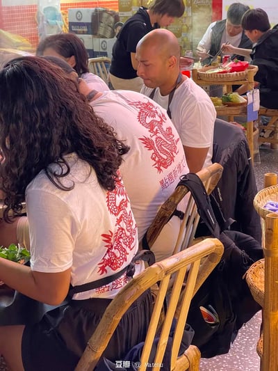 这是一处充满烟火气的中式火锅店场景，几位外国友人坐在竹制餐椅上享用火锅，其中两位的白色T恤背后印有醒目的红色中国龙图案，还配有部分可见的中文文字“點上選新增文”。店内还有其他食客正在就餐，竹制菜架上摆放着菜品，背景能看到BLUE GIRL的酒品广告，整体氛围热闹日常，展现了外国友人体验中式饮食文化的画面。