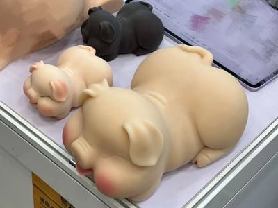 图片中展示了三个可爱的小猪摆件，两只米色和一只黑色，都呈现出熟睡的姿态。这些小猪造型圆润，有着闭着的眼睛和粉嫩的脸颊，显得十分治愈。它们被放置在一个白色的展示台上，背景中隐约可见一个平板电脑和部分人体部位。整个画面给人一种温馨可爱的感觉，适合作为家居装饰或礼品。