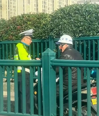 这是一张国内城市街头的实拍照片，画面被绿色的道路护栏分隔，左侧是身着荧光黄反光警服、佩戴制式警帽的正式执勤交警，右侧是一名坐在电动车上的男性驾驶人，该驾驶人头部佩戴的普通白色安全帽上手写有“交警”二字，疑似试图冒充交警逃避电动车交通规则检查，被真交警当场拦下，两人正隔着护栏面对面交流，背景可见道路绿化带和高层居民楼。