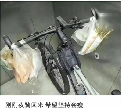 图片展示了一辆停在墙角的自行车，车把左右两侧各挂着一个白色塑料袋，左侧袋子装有疑似蔬菜的物品，右侧袋子内有细长状物品。自行车车架上配有一个深色小包，背景为灰色墙壁。图片底部文字“刚刚夜骑回来 希望坚持会瘦”表明这是用户夜骑后的场景，透露出通过坚持骑行实现减肥的期待。