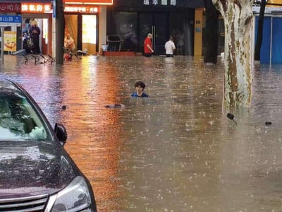 这是一张暴雨后的城市内涝现场实拍图，街道被浑浊的积水完全淹没，积水深度几乎没过画面中央男子的胸口，他在积水中艰难行进；画面左侧有一辆黑色轿车，车身大半被积水覆盖，车窗上还沾着雨点；背景处的商铺（瑞幸咖啡、哈尔滨私房菜等）门前也积满了水，有市民在店门口驻足观望，雨点仍在不断砸向水面，整个场景直观展现了城市遭遇暴雨内涝后的狼狈状况，传递出极端天气下出行的无奈。