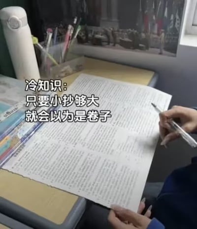 图片展示了一位学生在书桌前，在写一张大小如真正考试卷子般的大号小抄，桌面上有水杯、文具和学习材料。图片的配文：“冷知识：只要小抄够大就会以为是卷子”，用幽默方式调侃了考试的小抄和“卷子”的关系，具有双关与讽刺意味。“卷子”不仅指考试试卷，也暗含“内卷”现象，突出校园文化和学生群体中对学业压力的自嘲。