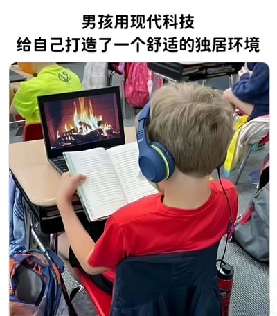 图片中展示了一个男孩在教室环境中，戴着蓝色耳机，面前的笔记本电脑屏幕上正播放着熊熊燃烧的壁炉视频，他手中还拿着一本打开的笔记本。图片上方配有文字“男孩用现代科技给自己打造了一个舒适的独居环境”，通过反差幽默的方式，调侃男孩在课堂上利用电子设备模拟舒适居家氛围的有趣场景。