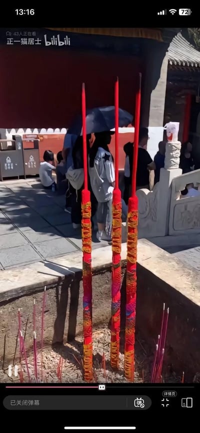 这是一张哔哩哔哩（B站）视频的竖屏截图，画面核心是寺庙的上香区域：三根装饰华丽的巨型供香插在香灰炉中，香身以红底搭配黄色彩纹与渐变彩饰，炉内还散落着不少已点燃或燃尽的细小红香，炉底堆积着香灰与残香。背景是典型的中式寺庙场景，红墙灰瓦搭配石质栏板，多名游客在此停留，有人撑着黑色遮阳伞，有人坐着休息，整体场景还原了寺庙中游客祈福上香的日常，氛围庄重又充满生活气息，展现了中式民俗里的上香祈福文化。