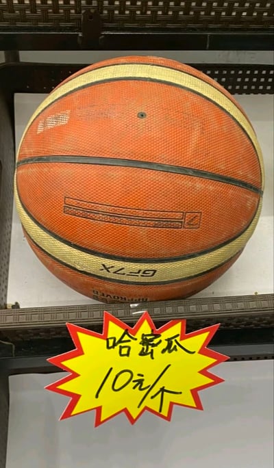 这是一张实拍的静态图片，画面中深色货架的层板上放置着一颗使用过的橙黄色标准篮球，篮球带有米黄色和黑色的条纹，球身印有倒过来的“GF7X”标识。篮球下方的层板边缘贴着一张黄底红边的爆炸形促销价格标签，标签上手写着“哈密瓜 10元/个”，显然是价格标签被错放到了篮球下方，形成了把篮球标注为哈密瓜售卖的错位趣味效果。