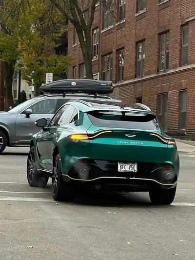 图片中，一辆绿色的Aston Martin SUV停在街头，装有车顶箱，最引人注目的是它的定制车牌“KFC V50”。这车牌让人联想到快餐品牌KFC，搭配豪车，形成了有趣的反差和幽默感，同时又可能带有车型代号的双关含义，容易引起路人的好奇和网友的玩梗。整体画面既有豪车范儿，又很接地气，堪称街头一景。
