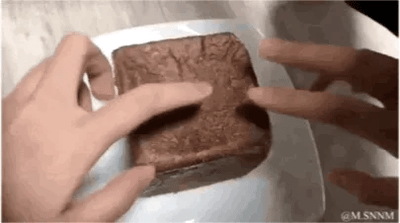 这是一段动态GIF，画面中一双手在白色盘子上方，缓慢地掰开一块外表呈巧克力色的面包状食物，随着双手的动作，面包内部浓稠丝滑的巧克力流心缓缓流淌出来，呈现出极具诱惑力的爆浆效果，整个过程视觉上十分治愈，能给人带来满足的美食体验，没有双关、谐音梗或暗示类内容。