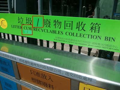 图片展示了一个绿色的垃圾/废物回收箱，箱体上方有中英文标识，中文为“垃圾/废物回收箱”，英文标注为“LITTER CUM RECYCLABLES COLLECTION BIN”。其中“CUM”一词被红色方框重点圈出，显得格外突兀。标识下方还有补充说明文字：“如發現此回收箱滿溢或有任何投訴，請致電1823電話中心 If this bin is full or you have any complaint, please contact 1823 Call Centre”。背景中可见建筑物入口和一名站立的行人，整体场景为户外公共场所。

图片中的“CUM”在英文中有粗俗含义，此处明显是标识制作时的错误，可能是想表达“AND”或误植了分隔符号，红框标注意在突出这一尴尬失误。