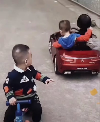 室外水泥地面场景中，一名穿着黑色带彩色图案毛衣的小男孩正骑在蓝色三轮车上，伸出手指向不远处一辆红色玩具车。玩具车后部有奔驰标志和“AMG”字样，车内坐着另一名穿蓝色连帽衫（袖子为红色）的小男孩，驾驶座旁的乘客位上放着一个黑色球状物体（疑似气球或头盔）。骑三轮车的男孩扭头注视着玩具车，表情中带着好奇与向往，整体画面展现了儿童间纯真的互动与童年玩乐场景。