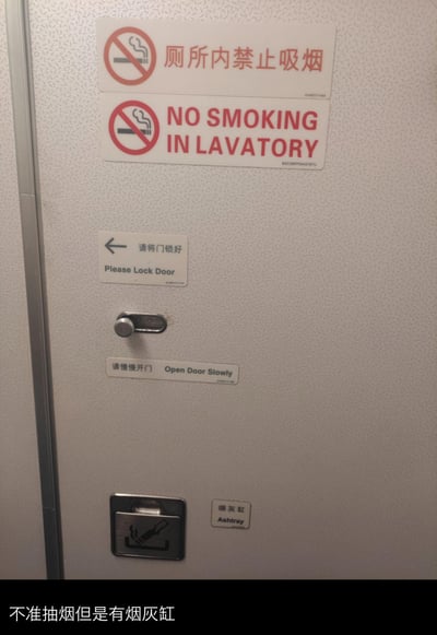 这是一张民航客机卫生间的实拍图，墙面上张贴了多组中英双语提示标识：最上方是两块醒目的禁止吸烟标识，分别标注“厕所内禁止吸烟”和“NO SMOKING IN LAVATORY”；下方依次是“请将门锁好 Please Lock Door”的锁门提示、门锁装置、“请慢慢开门 Open Door Slowly”的开门提示，最底部则设置了一个烟灰缸装置，旁边配有“烟灰缸 Ashtray”的标识。图片底部配文“不准抽烟但是有烟灰缸”，点出了禁烟提示与烟灰缸共存的矛盾感。实际上这种设置是民航安全规范的要求：全面禁烟后保留烟灰缸，是为了处理乘客违规吸烟后的烟蒂，避免烟蒂随意丢弃引发安全隐患。
