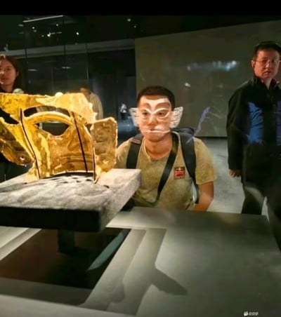 这是一张在博物馆展厅内拍摄的趣味照片，画面中的游客坐在展柜前，正对着陈列的金色面具，巧妙的拍摄角度使得金面具与他的头部重合，营造出游客像是戴上了金面具的错位幽默效果。这种场景常用于朋友间互动、文博热潮下的展馆打卡照，非常适合表达轻松和有趣的氛围。图片属于经典错位合影梗，既突出展品亮点，又带来搞笑趣味。