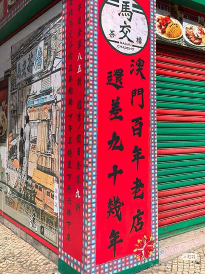 这张是实拍澳门本地餐饮店铺“马交茶档”的门店外墙宣传，图片整体为街头实景拍摄，店铺转角立柱设置了一块醒目的红底黑字竖幅宣传广告：顶部是圆形店铺标识，标注店名“馬交茶檔”与英文“MACAU CHADONG”，店名“马交”是“Macau（澳门）”的音译，下方竖写大字宣传语“澳門百年老店 還差九十幾年”，玩了趣味文字梗：该店实际开业仅3年，以“距离百年老店还差九十几年”的幽默反差表述，表明做百年老店的长期经营目标，十分有趣。竖幅侧边的立柱另一面标注了店铺的促销折扣信息：庆祝开店三周年，平日全场八五折，周末节假日全场九折，下午茶时段下单适用该优惠。背景墙面印有复古风格的澳门旧街景喷绘，画中有背对镜头的行人，还原老澳门街肆氛围，画面右侧是红绿相间的店铺卷闸门，整体是复古又充满趣味的实体店宣传布置。