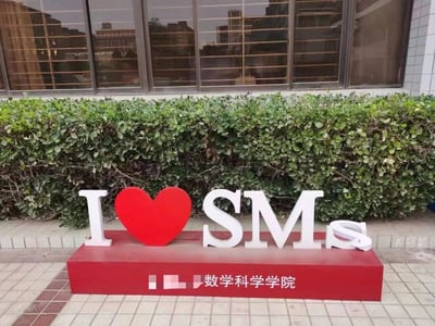 图片展示了一个位于校园内的标牌，标牌主体为红色底座，上面有白色的‘I’字母、红色的心形符号和白色的‘SMS’字母，组合成‘I ❤️ SMS’的图案。底座下方印有‘数学科学学院’字样。标牌后方是修剪整齐的绿色灌木丛，背景为一栋带有大面积玻璃窗的建筑，窗户反射出周围的楼宇景象。整体场景给人一种校园环境中的温馨与学术氛围。